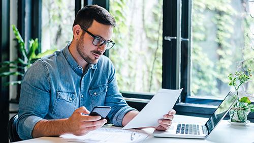 Man wearing glasses looking at paper while holding cellphone